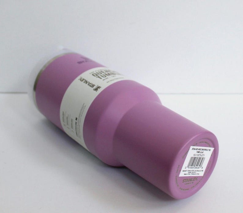 May include: A lavender-colored Stanley Quencher Tumbler with a clear plastic lid. The tumbler has a cylindrical body with a tapered base. A white label wraps around the body with product information. The tumbler is designed for cold beverages.