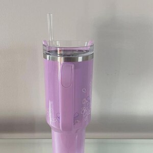 May include: A lavender insulated tumbler with a clear lid and a clear straw. The tumbler has a handle and is decorated with a floral pattern. The tumbler is designed to hold cold beverages.