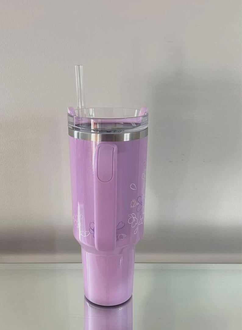 May include: A lavender insulated tumbler with a clear lid and a clear straw. The tumbler has a handle and is decorated with a floral pattern. The tumbler is designed to hold cold beverages.