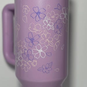 May include: A lavender insulated tumbler with a handle and a clear lid. The tumbler is decorated with a floral design in white and purple. The tumbler is designed to hold cold or hot beverages.