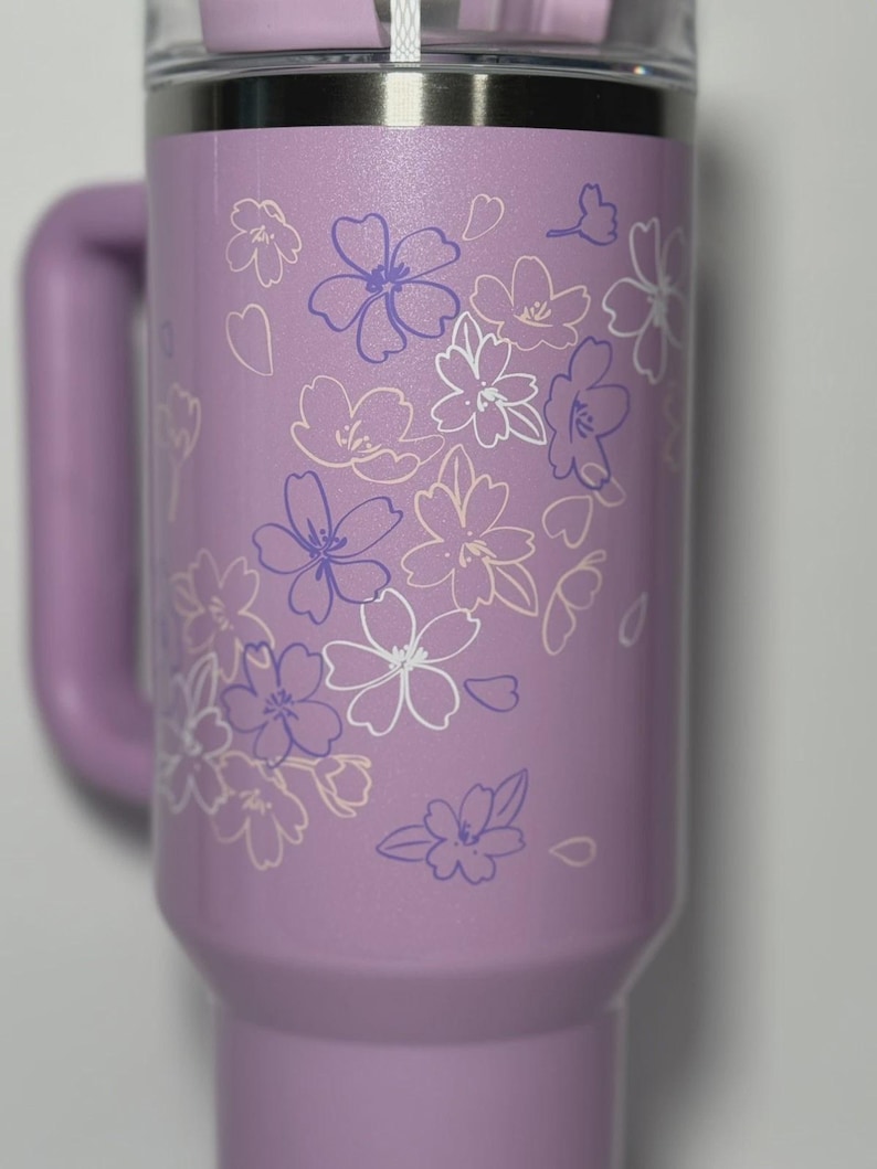 May include: A lavender insulated tumbler with a handle and a clear lid. The tumbler is decorated with a floral design in white and purple. The tumbler is designed to hold cold or hot beverages.