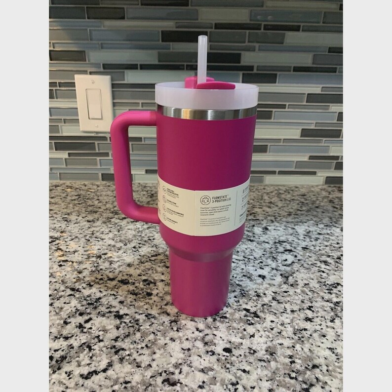 Puede incluir: Un vaso t&eacute;rmico magenta con asa, tapa blanca y pajita. El vaso tiene un cuerpo cil&iacute;ndrico y un asa lateral. Una etiqueta blanca envuelve el cuerpo.