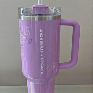 May include: A lavender Stanley + Starbucks tumbler with a handle and a clear straw. The tumbler features a floral design and the brand name. The lid is clear. The tumbler is designed to hold a large quantity of liquid.
