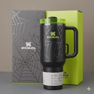 May include: Black Stanley tumbler with a spiderweb design and a green lid. The tumbler has a black handle and the Stanley logo in green. The tumbler is next to a gray box with a spiderweb design and a green box.