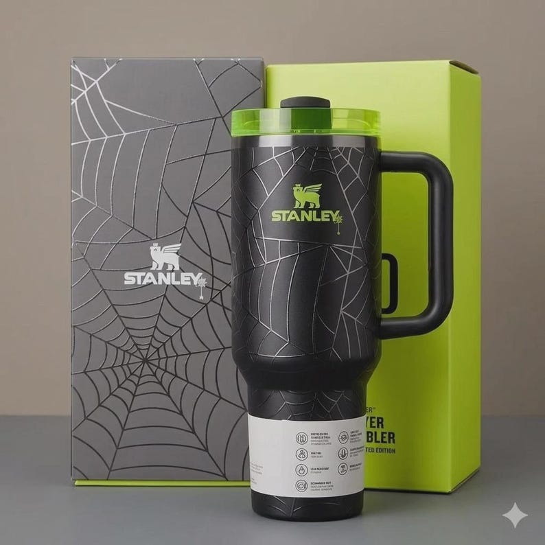 May include: Black Stanley tumbler with a spiderweb design and a green lid. The tumbler has a black handle and the Stanley logo in green. The tumbler is next to a gray box with a spiderweb design and a green box.