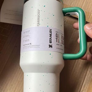May include: A white Stanley Quencher tumbler with a green handle and accents. The tumbler has a speckled design and a Starbucks logo. It holds 40 oz and is designed for all-day hydration.