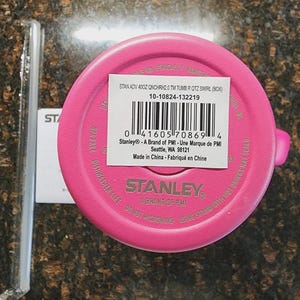 May include: A bright pink Stanley tumbler lid with a white label and barcode. The label includes the text "STAN ADV 400Z QNCHRH2.0 TM TUMB R QTZ SWIRL (BOX)" and "Stanley - A Brand of PMI". A clear plastic straw is visible.