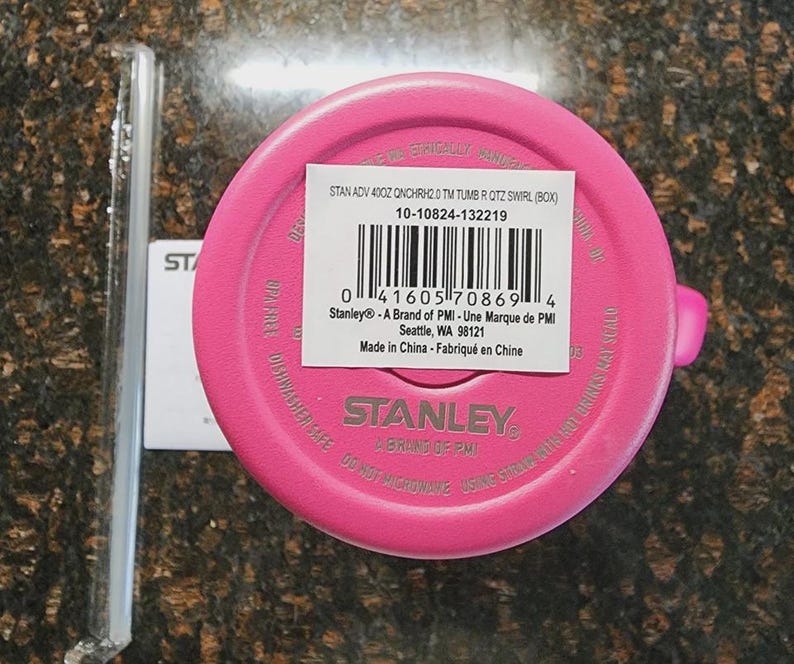 May include: A bright pink Stanley tumbler lid with a white label and barcode. The label includes the text "STAN ADV 400Z QNCHRH2.0 TM TUMB R QTZ SWIRL (BOX)" and "Stanley - A Brand of PMI". A clear plastic straw is visible.