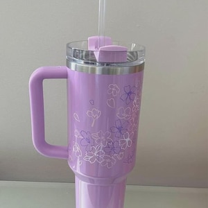 May include: A lavender insulated tumbler with a handle, clear lid, and straw. The tumbler features a floral design in white and purple. The handle, lid, and straw are the same lavender color as the tumbler.