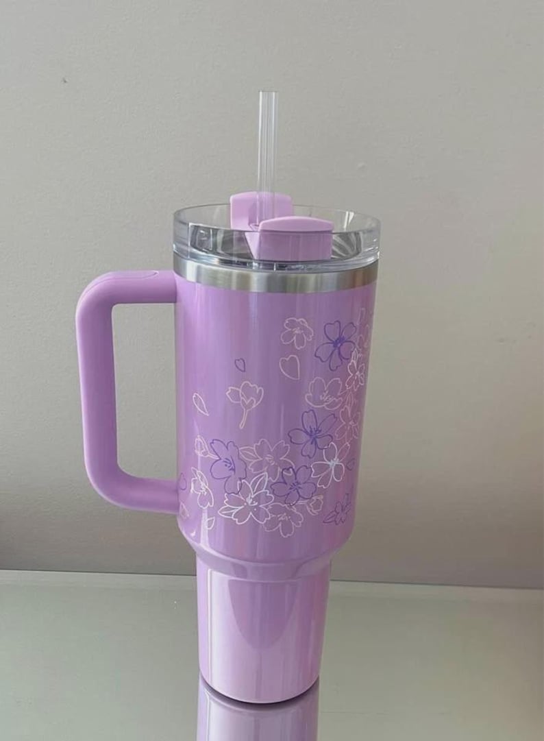 May include: A lavender insulated tumbler with a handle, clear lid, and straw. The tumbler features a floral design in white and purple. The handle, lid, and straw are the same lavender color as the tumbler.