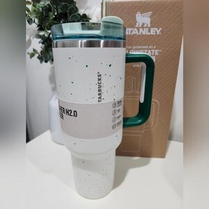 May include: A white and stainless steel tumbler with a green handle and lid. The tumbler has a speckled design with dark teal accents and the text "STARBUCKS" and "QUENCHER H2.0" printed on the side. The tumbler is approximately 10 inches tall.