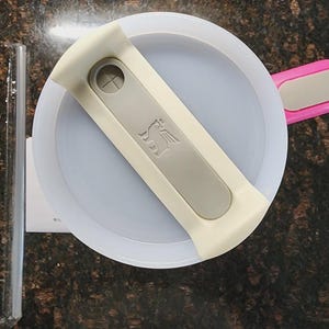 May include: Overhead view of a white and gray Stanley brand food container lid with a gray handle and a pink handle. A clear glass tube and a white paper are on the left side of the image.