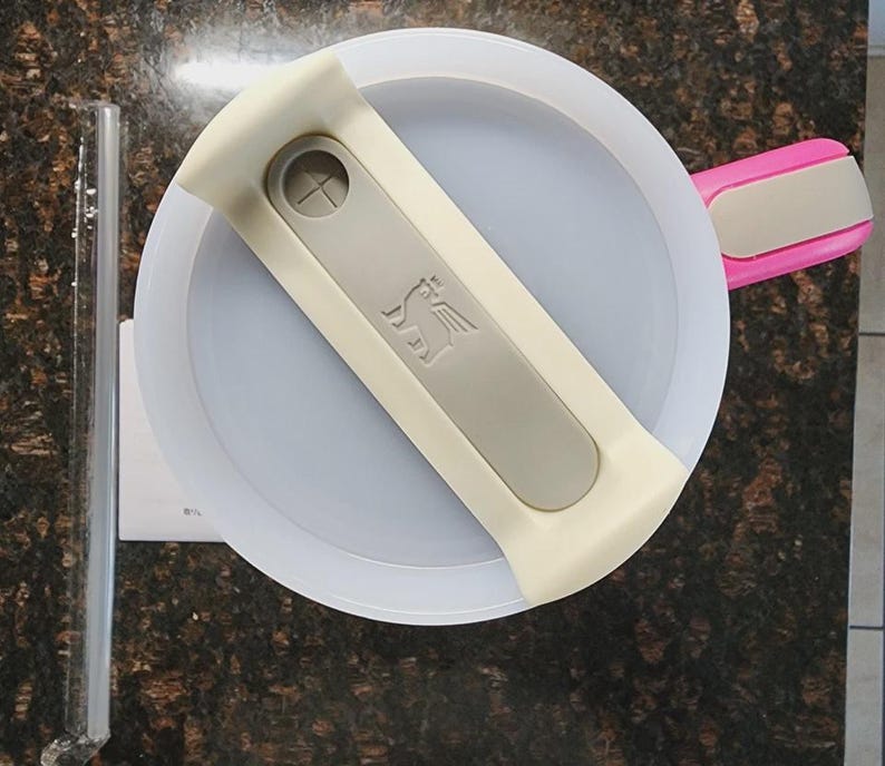 May include: Overhead view of a white and gray Stanley brand food container lid with a gray handle and a pink handle. A clear glass tube and a white paper are on the left side of the image.
