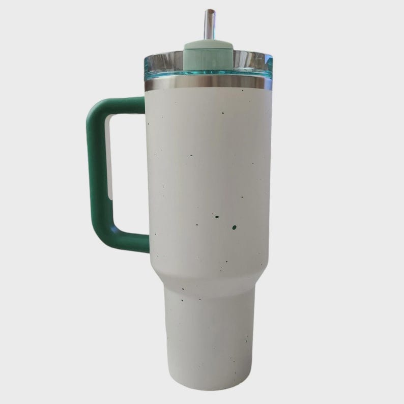 May include: A white insulated tumbler with a green handle and lid. The tumbler has a stainless steel rim and a clear lid with a green top and a straw. The tumbler is designed to keep beverages cold or hot.