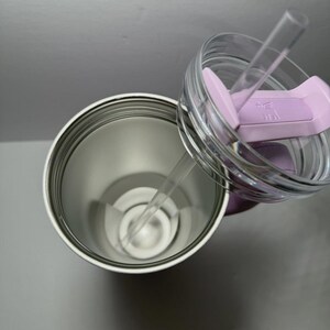 May include: A stainless steel tumbler with a clear lid and a light purple slider. The lid has a clear straw. The tumbler is open, revealing the interior. The background is a light gray.