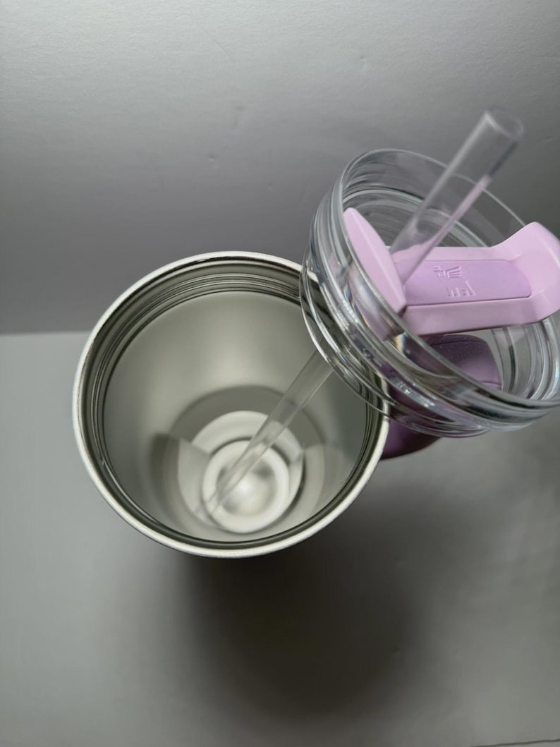 May include: A stainless steel tumbler with a clear lid and a light purple slider. The lid has a clear straw. The tumbler is open, revealing the interior. The background is a light gray.