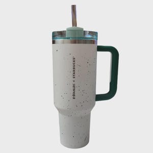May include: A white and speckled Stanley + Starbucks tumbler with a green handle and lid. The tumbler has a stainless steel straw and a clear lid. The tumbler is designed to keep drinks cold or hot.