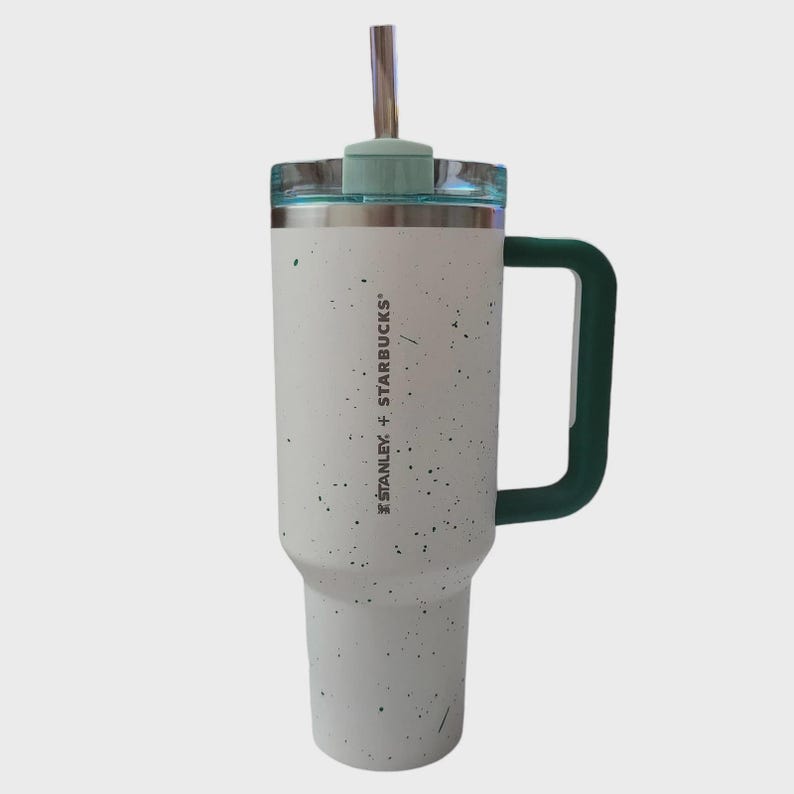 May include: A white and speckled Stanley + Starbucks tumbler with a green handle and lid. The tumbler has a stainless steel straw and a clear lid. The tumbler is designed to keep drinks cold or hot.
