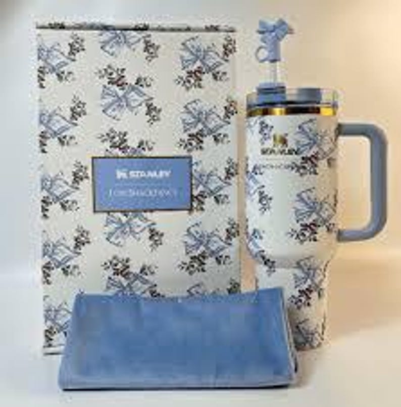 May include: A Stanley tumbler with a floral pattern, a matching box, and a blue cloth. The tumbler has a handle and a straw. The box has a blue label that reads "STANLEY FOR SHACK ATTACK". The tumbler is approximately 9 inches tall.