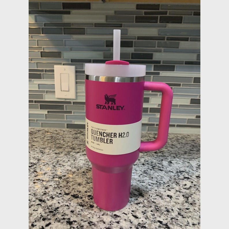 Puede incluir: Un vaso Stanley Quencher H2.0 magenta con tapa y asa blancas. El vaso tiene una pajita blanca y una etiqueta blanca con texto negro "QUENCHER H2.0 TUMBLER". El logotipo de Stanley es visible.