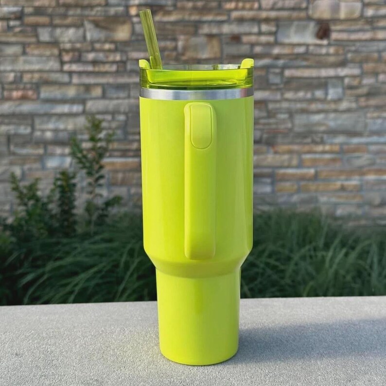 May include: A bright neon green insulated tumbler with a handle and a clear lid. The tumbler has a matching green straw and a silver rim. The design is sleek and modern, perfect for keeping drinks cold or hot.