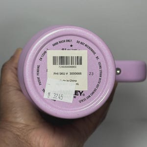 May include: A light purple insulated tumbler with a handle. The bottom of the tumbler has a sticker with a barcode and text. The text on the tumbler includes "HAND WASH ONLY" and "DO NOT MICROWAVE".