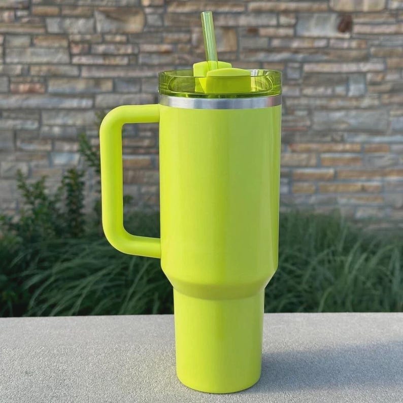 May include: A bright neon green insulated tumbler with a handle, lid, and straw. The tumbler has a silver rim and a clear lid. The handle is a matching neon green color. The straw is also neon green.