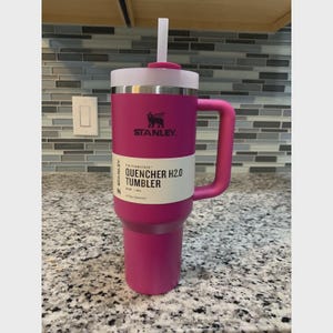 Puede incluir: Un vaso Stanley Quencher H2.0 magenta con tapa y pajita blancas. El vaso tiene un asa y el logotipo de Stanley. El texto "QUENCHER H2.0 TUMBLER" est&aacute; impreso en el lateral. Dise&ntilde;ado para la hidrataci&oacute;n durante todo el d&iacute;a.