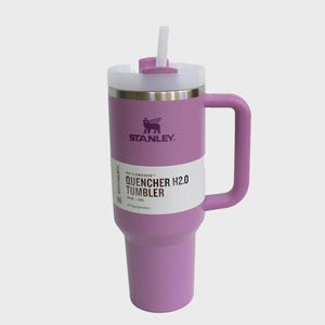 May include: A lavender Stanley Quencher H2.0 Tumbler with a white straw and handle. The tumbler has a white label with black text that reads "QUENCHER H2.0 TUMBLER". The Stanley logo is visible on the front of the tumbler.