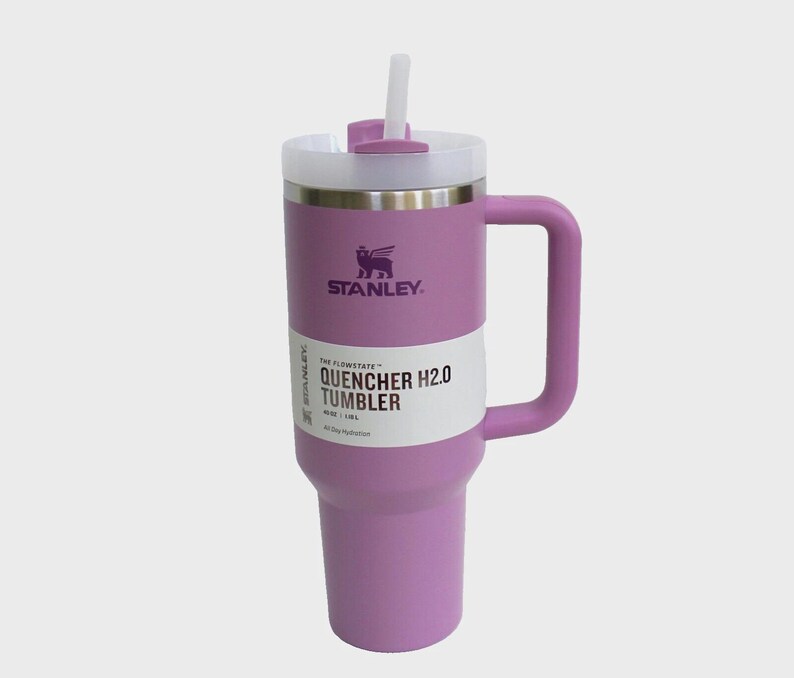 May include: A lavender Stanley Quencher H2.0 Tumbler with a white straw and handle. The tumbler has a white label with black text that reads "QUENCHER H2.0 TUMBLER". The Stanley logo is visible on the front of the tumbler.