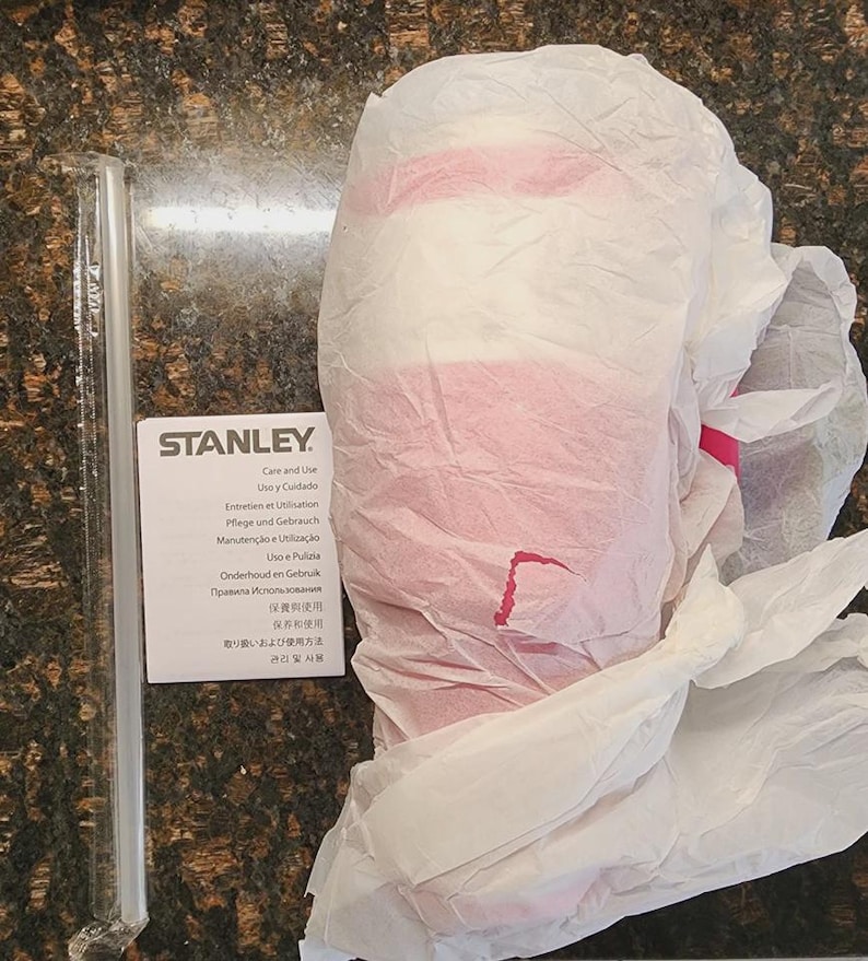 May include: A Stanley product, wrapped in white tissue paper with pink accents. A clear straw and a care instruction card are visible. The card includes the Stanley logo and text in multiple languages.