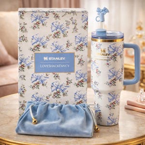 May include: A Stanley Loveshackfancy tumbler set with a matching box and velvet pouch. The tumbler, box, and pouch feature a white background with blue bows and floral patterns. The tumbler has a blue handle and straw with a bow accent.