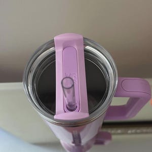 May include: A light purple tumbler with a clear lid and a matching straw. The tumbler has a handle and a logo on the side. The lid has a sliding closure for the straw. The tumbler is designed to keep drinks cold or hot.