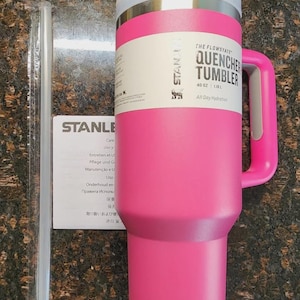 May include: A pink Stanley Quencher Tumbler with a handle, a clear straw, and a white and silver lid. The tumbler has the text "THE FLOWSTATE QUENCHER TUMBLER 40 OZ" printed on it. A care instruction sheet is also visible.
