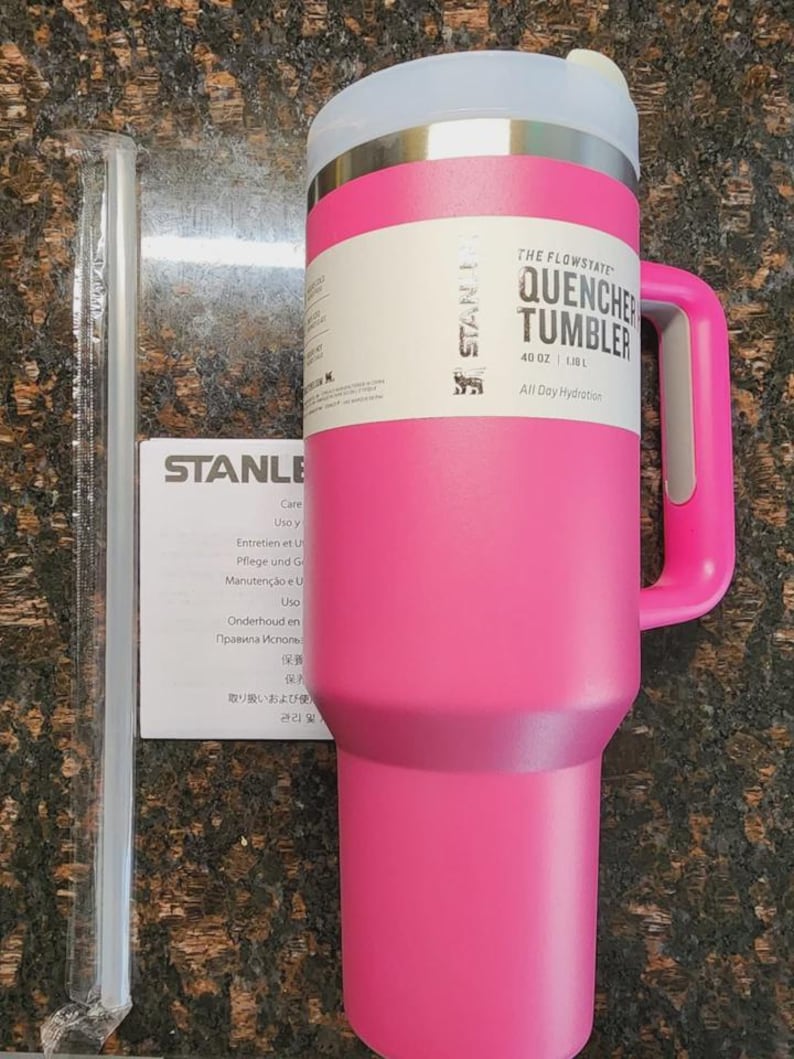 May include: A pink Stanley Quencher Tumbler with a handle, a clear straw, and a white and silver lid. The tumbler has the text "THE FLOWSTATE QUENCHER TUMBLER 40 OZ" printed on it. A care instruction sheet is also visible.