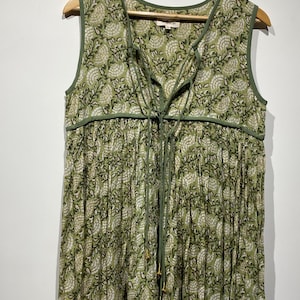 May include: Sleeveless green dress with a floral pattern. The dress features a V-neckline with a tie closure and green trim. The fabric has a pleated design, and the dress is hanging on a wooden hanger.