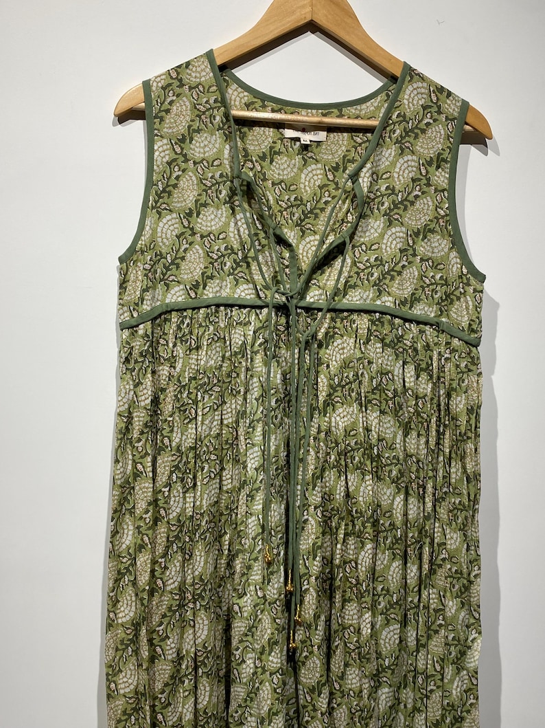 May include: Sleeveless green dress with a floral pattern. The dress features a V-neckline with a tie closure and green trim. The fabric has a pleated design, and the dress is hanging on a wooden hanger.
