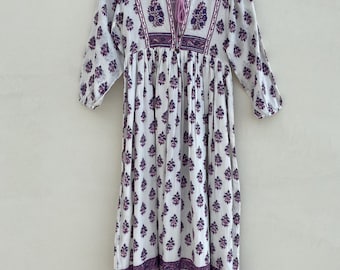 May include: A white dress with a purple floral pattern and three-quarter sleeves. The dress features a V-neckline with a drawstring and is hanging on a wooden hanger. The floral design is repeated throughout the dress, with a border at the hem.