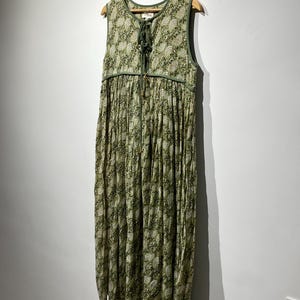 May include: A sleeveless, floor-length dress with a green and white floral pattern. The dress features a V-neckline with a drawstring closure and a gathered skirt. The dress is hanging on a wooden hanger against a white wall.