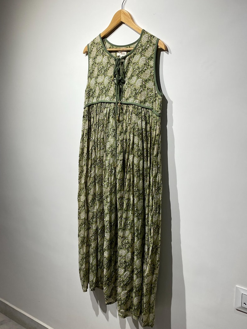 May include: A sleeveless, floor-length dress with a green and white floral pattern. The dress features a V-neckline with a drawstring closure and a gathered skirt. The dress is hanging on a wooden hanger against a white wall.