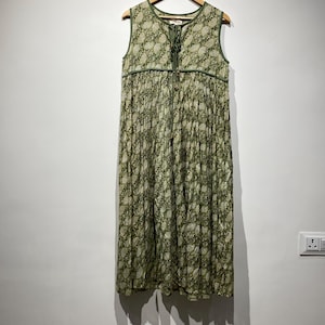 May include: A sleeveless, floor-length dress with a green and white floral pattern. The dress is hanging on a wooden hanger against a white wall. It features a V-neckline with a tie closure and green trim.