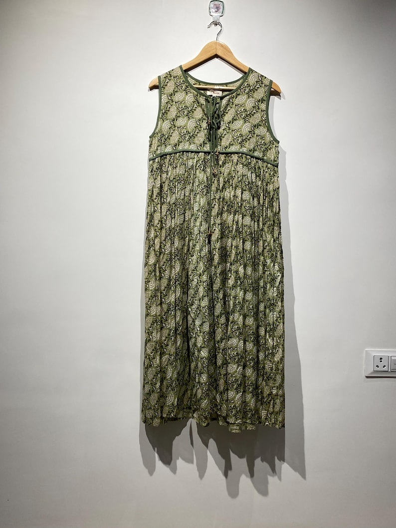 May include: A sleeveless, floor-length dress with a green and white floral pattern. The dress is hanging on a wooden hanger against a white wall. It features a V-neckline with a tie closure and green trim.