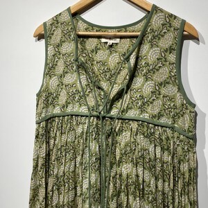 May include: A sleeveless green dress with a floral pattern and a lace-up neckline. The dress is hanging on a wooden hanger. The fabric has a white and green floral design. The dress has a gathered skirt.