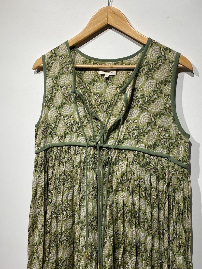 May include: A sleeveless green dress with a floral pattern and a lace-up neckline. The dress is hanging on a wooden hanger. The fabric has a white and green floral design. The dress has a gathered skirt.