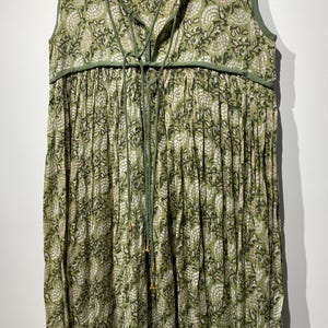 May include: Sleeveless green dress with a floral pattern in white and green. The dress features a V-neckline with a drawstring and a gathered skirt. The trim and drawstring are a darker shade of green.