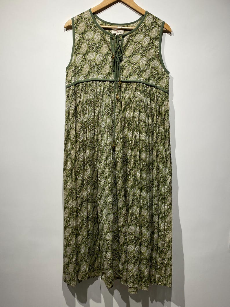 May include: Sleeveless green dress with a floral pattern. The dress has a round neckline, a drawstring detail at the chest, and a pleated skirt. The dress is hanging on a wooden hanger.