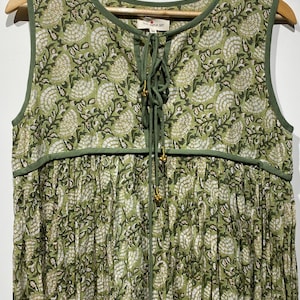 May include: A sleeveless green dress with a floral pattern. The dress has a round neckline and a drawstring closure with gold-colored beads. The fabric is a light green color with a white and dark green floral design. The dress is hanging on a wooden hanger.