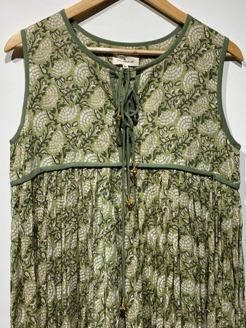 May include: A sleeveless green dress with a floral pattern. The dress has a round neckline and a drawstring closure with gold-colored beads. The fabric is a light green color with a white and dark green floral design. The dress is hanging on a wooden hanger.
