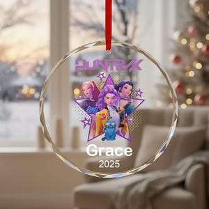 Peut inclure: Une boule de Noël ronde et transparente avec un ruban rouge. L'ornement présente un graphisme de trois personnages de dessins animés et le texte "HUNTIK", "Grace" et "2025". L'arrière-plan comprend un sapin de Noël et une fenêtre.