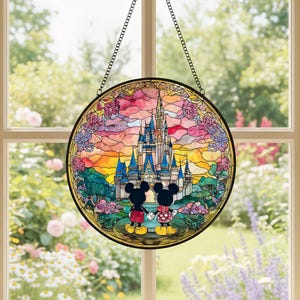 May include: A circular stained glass window depicting a castle scene with Mickey and Minnie Mouse. The artwork is framed in black and suspended by a chain. The background features a colourful sunset and floral details.